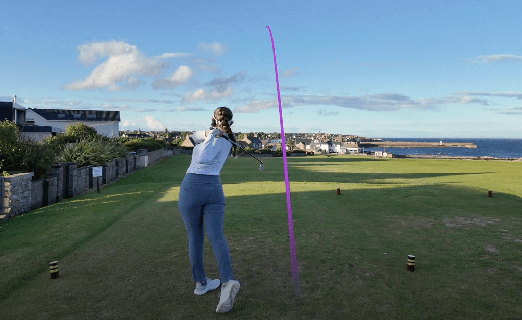 A woman hitting a tee shot on the final hole at Anstruther Golf Club, with a pink tracer showing the trajectory of the golf ball