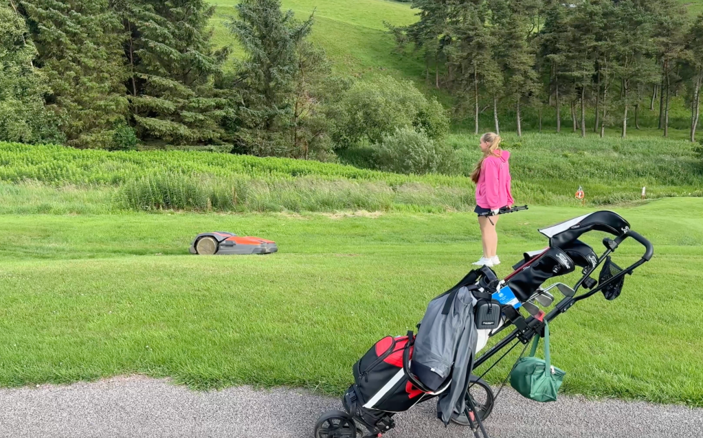 Georgina stands beside a Husqvarna CEORA robotic mower at Eastwood Twelve, with her golf bag in front of her.